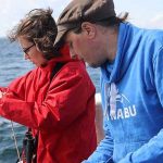 Eine Frau in NAJU-Kleidung und ein Mann in NABU-Kleidung lassen einen Behälter zur Wasserprobe in die Ostsee.